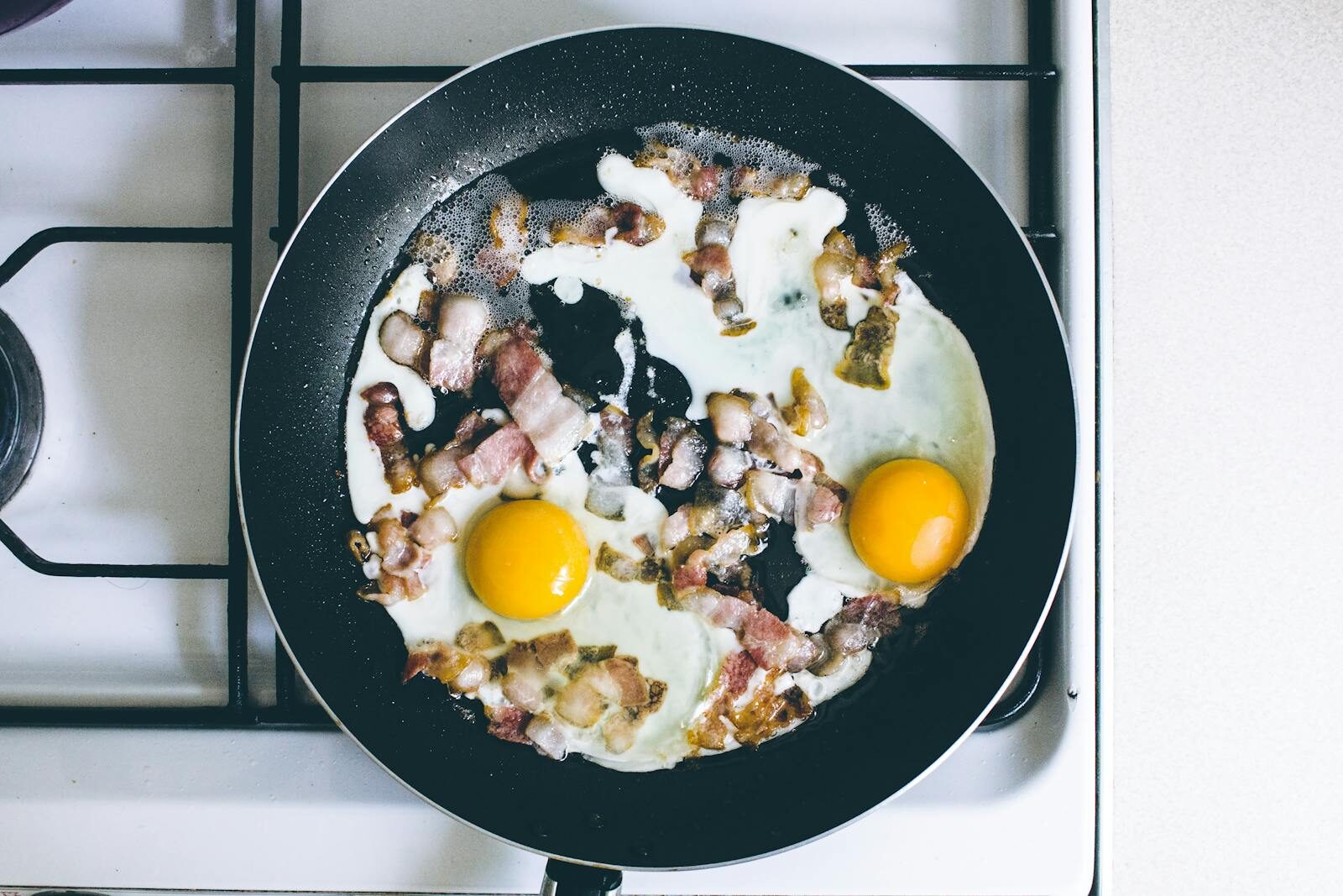 High-angle view of fried eggs and bacon sizzling in a skillet on a gas stove, perfect breakfast scene.