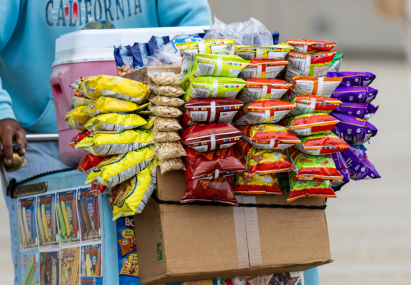 A street vendor's cart filled with a variety of colorful snack packets for sale outdoors.